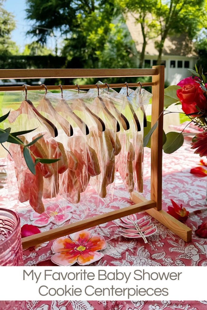 A wooden stand holds decorated cookies in clear bags, displayed as a centerpiece on a floral-patterned table at an outdoor baby shower.