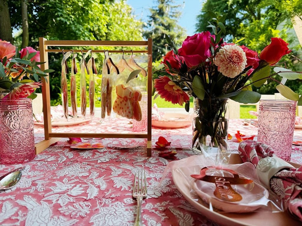 A pink floral tablecloth with a floral centerpiece, pink glassware, decorated cookies on a stand, and a plate with cookies and napkins set for an outdoor meal.