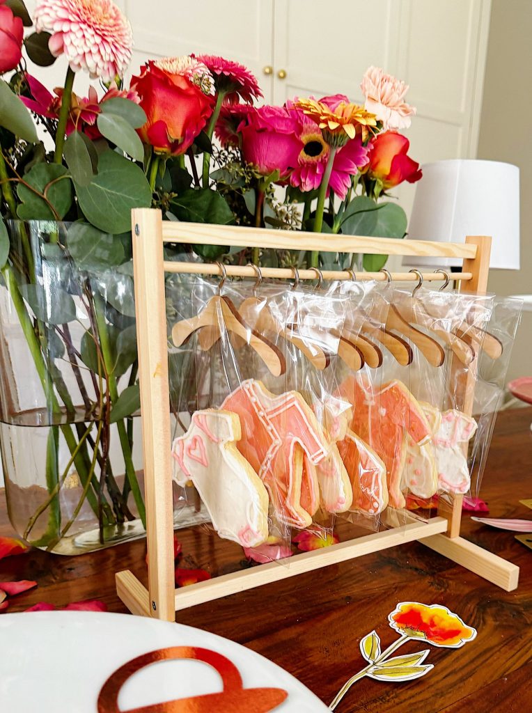 A wooden rack holds decorated cookies shaped like dresses in clear bags, displayed in front of a vase of colorful flowers on a table.