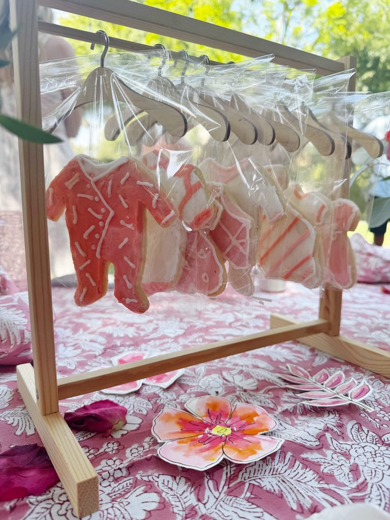 Decorated cookies shaped like baby onesies and hung on a wooden rack with mini hangers, each cookie wrapped in plastic, displayed on a floral tablecloth.