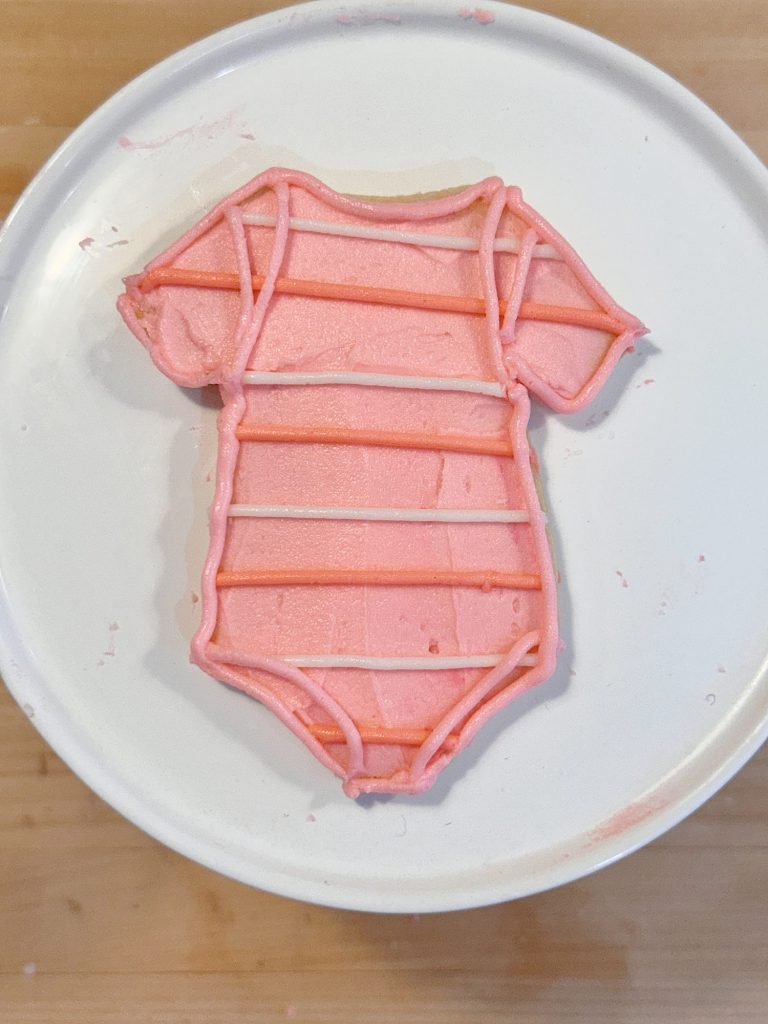 A cookie shaped like a baby onesie, decorated with pink icing and darker pink stripes, sits on a white plate.