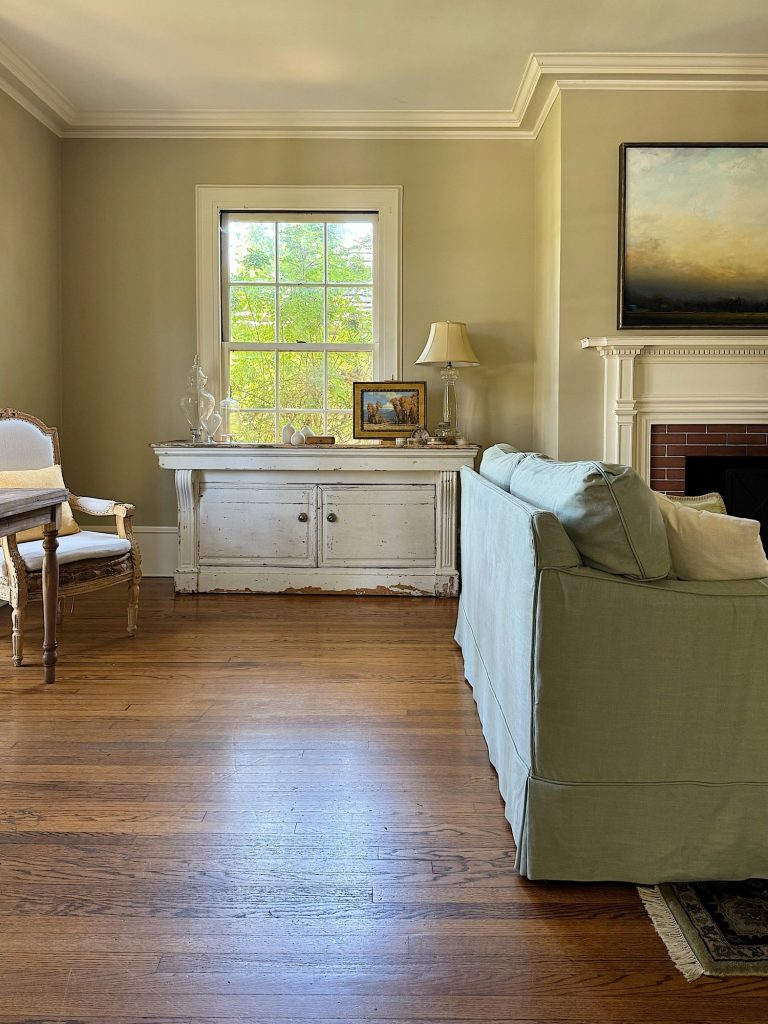 A living room with a wooden floor, light green sofa, vintage white console table, upholstered chair, table lamp, framed photo, and window letting in natural light.