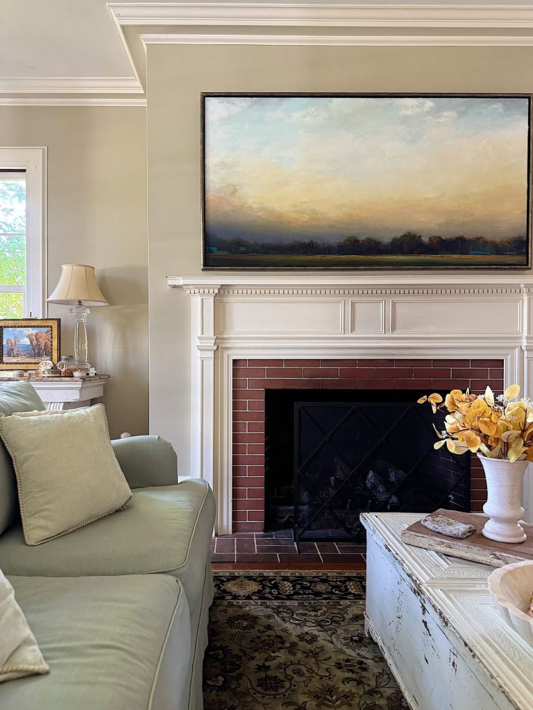 A living room with a light green sofa, white mantel fireplace, landscape painting above the mantel, and a coffee table with a vase of yellow flowers.