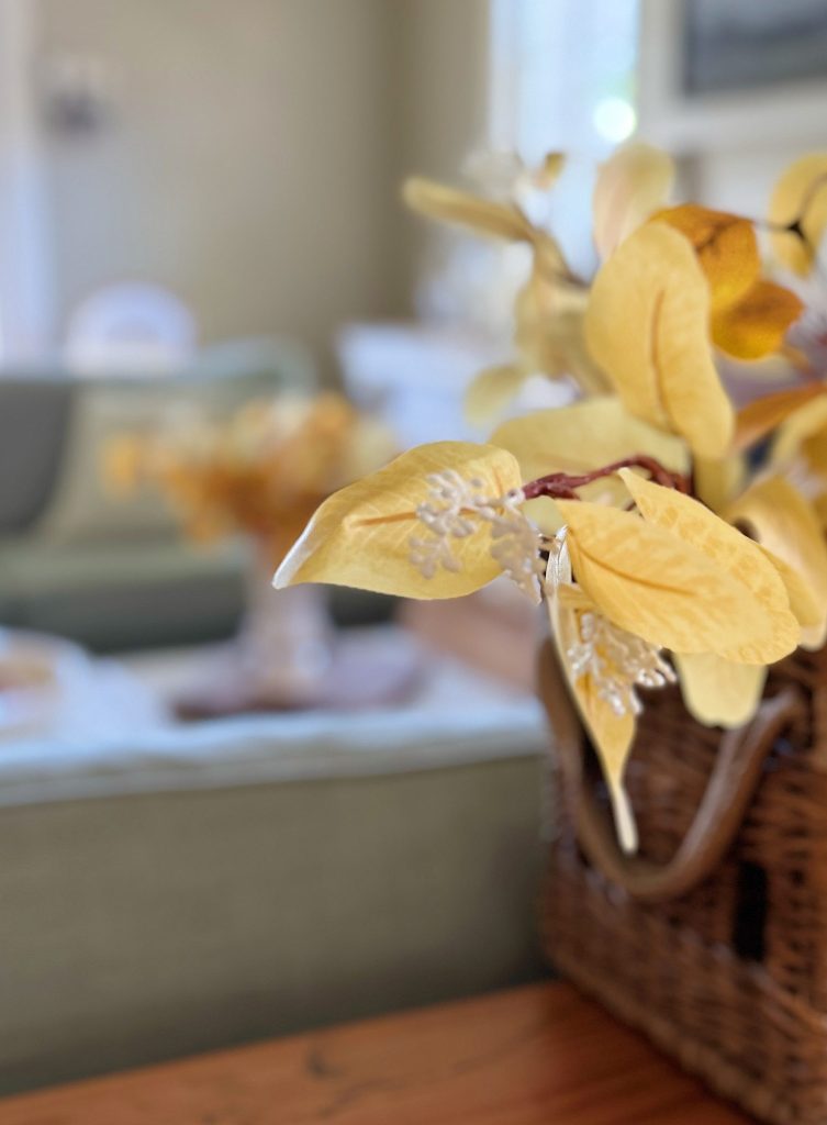 Close-up of yellow artificial flowers in a woven basket on a wooden table, with a blurred living room scene in the background.