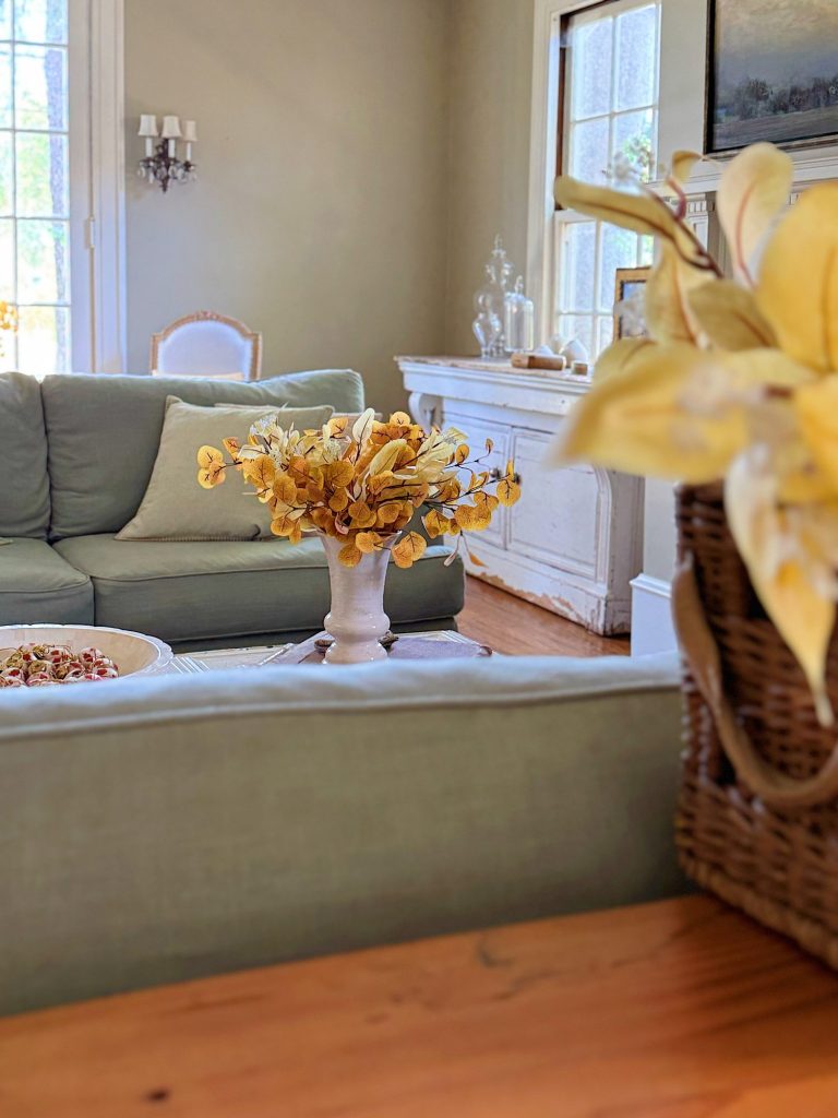 A living room with light green sofas, a white vase of yellow flowers on a table, a basket with yellow leaves, and sunlight streaming through large windows.