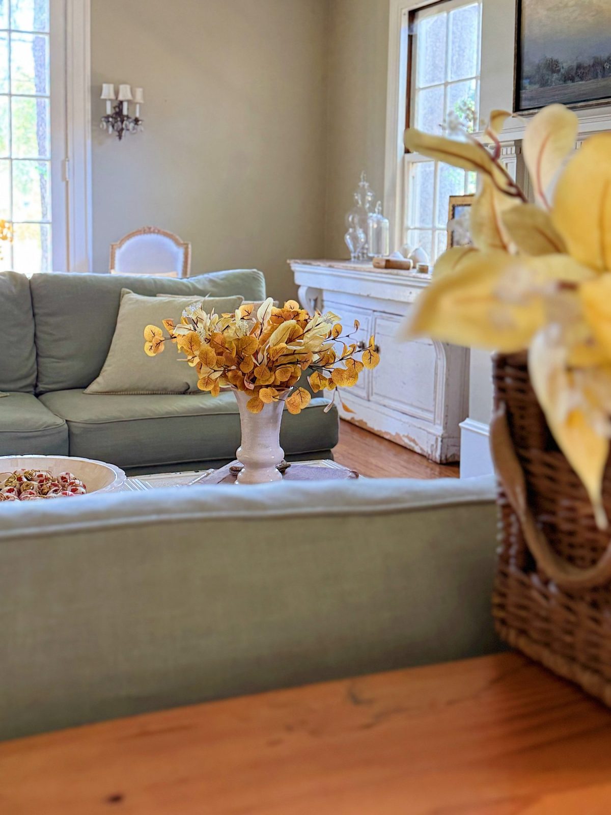 A living room with light green sofas, a white vase of yellow flowers on a table, a basket with yellow leaves, and sunlight streaming through large windows.