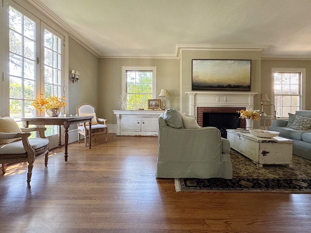 A bright living room with wooden floors, light-colored furniture, large windows, a fireplace, and a landscape painting above the mantel.