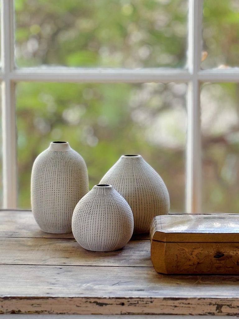 Three textured white ceramic vases of varying sizes and a rectangular wooden box are arranged on a wooden table in front of a window with a leafy green view.