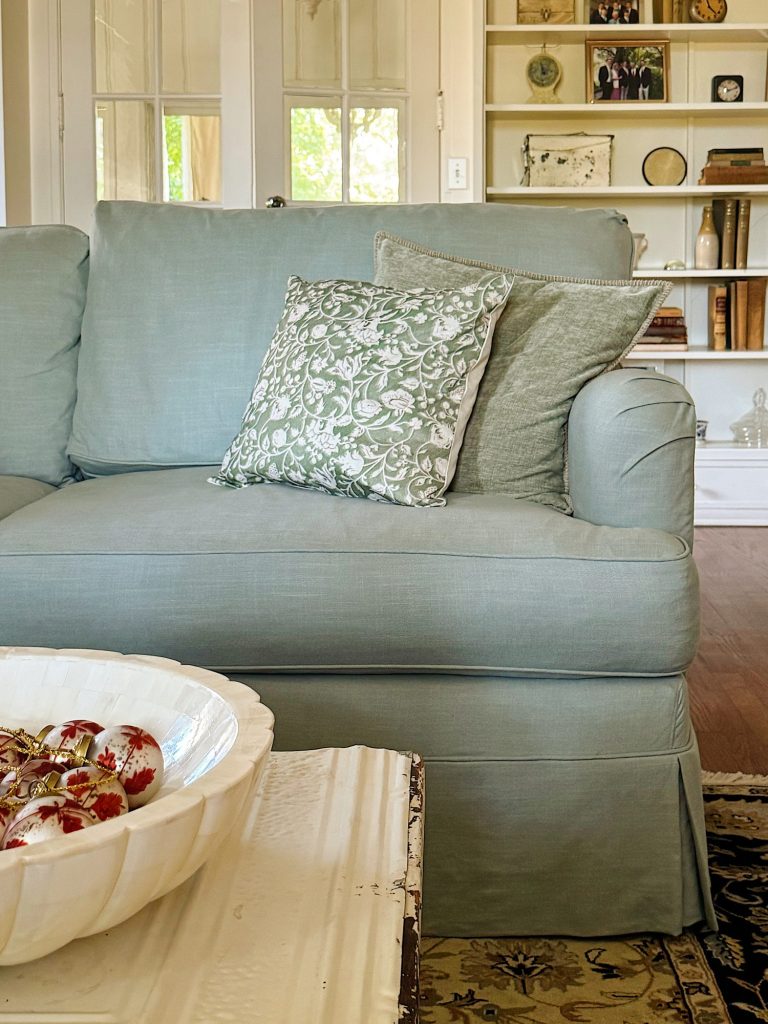 Light blue upholstered sofa with two patterned cushions sits in a living room with built-in shelves in the background and a decorative bowl on a white coffee table in the foreground.