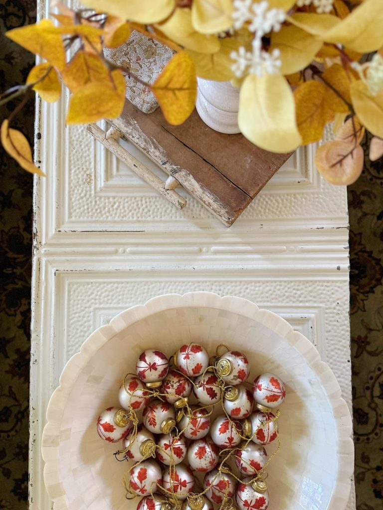 A white bowl filled with red and white maple leaf ornaments sits on a textured cream-colored table beside a wooden tray with a vase of yellow and white dried flowers.