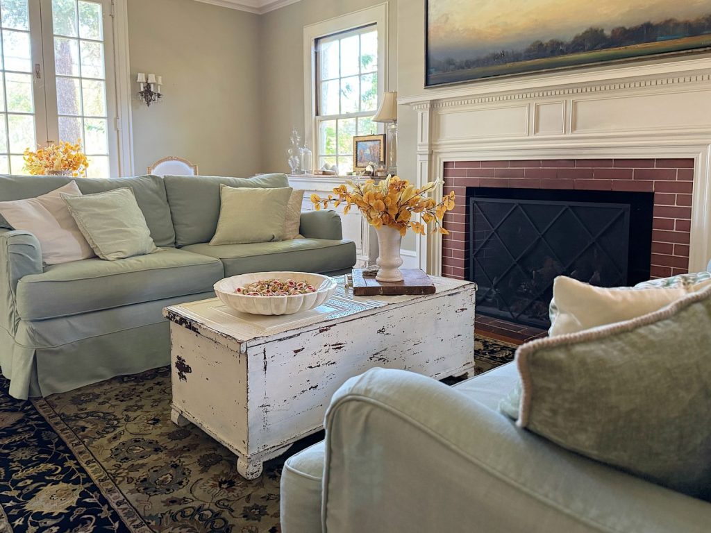 Light-filled living room with two pale green sofas, a distressed white coffee table, a decorative bowl, and a fireplace with a painting above the mantel.