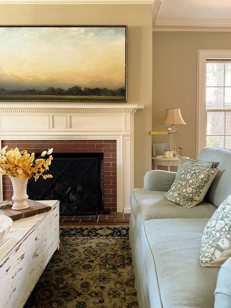 A living room with a light blue sofa, patterned pillows, a brick fireplace with a large landscape painting above, and a rustic white coffee table with a yellow floral arrangement.
