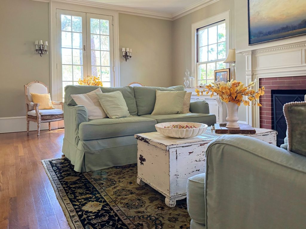 A living room with light green sofas, a white vintage coffee table, a fireplace, large windows, and a floral arrangement on the table.