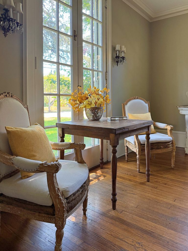 Two upholstered armchairs with yellow pillows flank a wooden table holding a basket of yellow flowers in front of tall French doors in a sunlit room with hardwood floors.