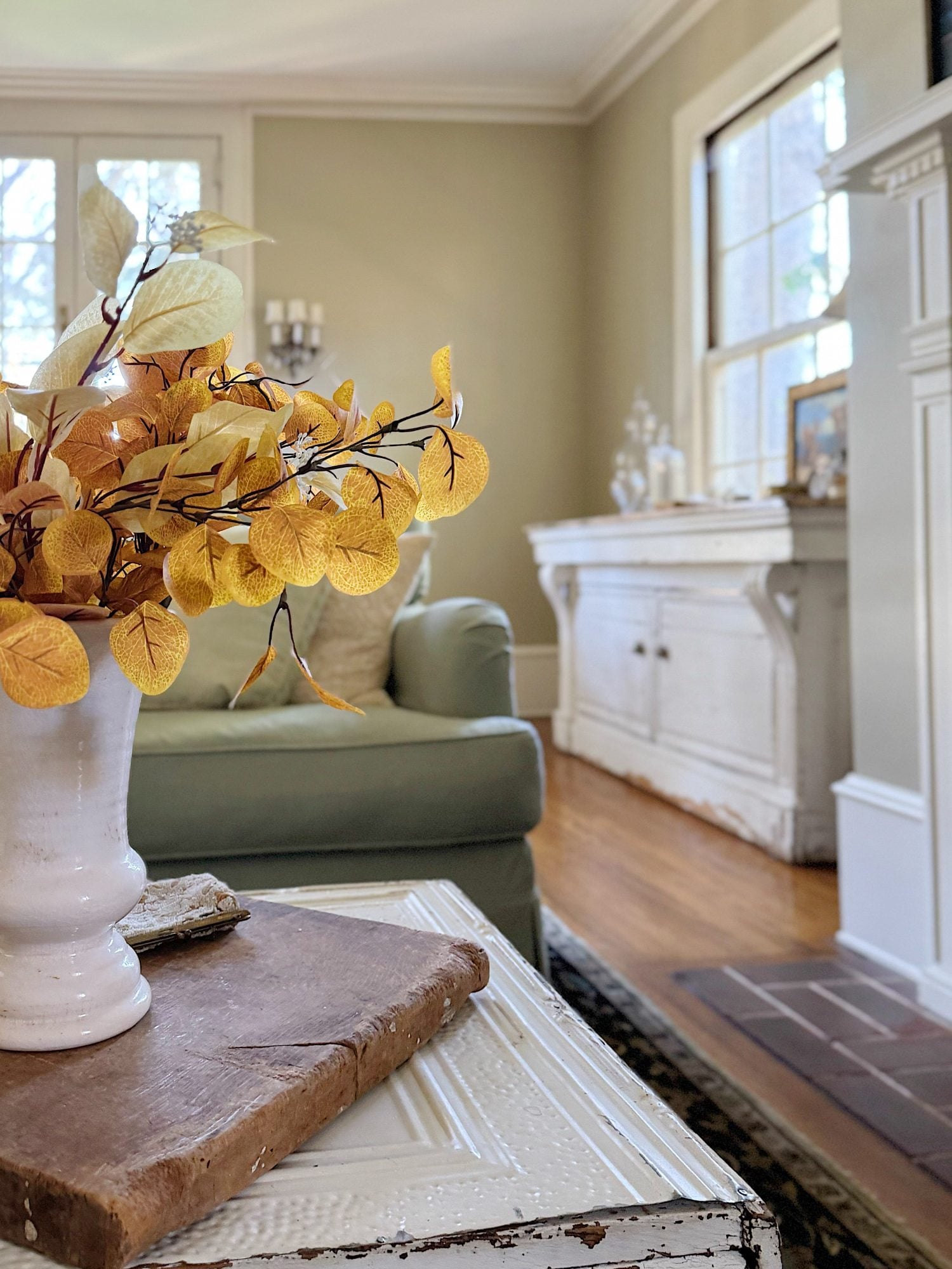 A white vase with yellow leaves sits on a table next to a book in a cozy living room with a sofa, white sideboard, and wooden floors.