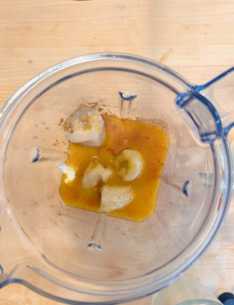 Blender with banana pieces, orange liquid, and some spices inside, viewed from above on a wooden surface.