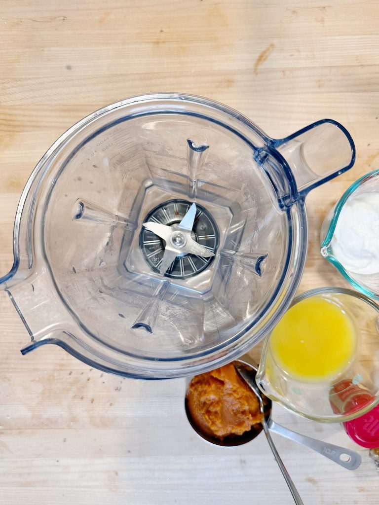 Top view of an empty blender surrounded by a measuring cup of orange liquid, another of thick white substance, and a metal cup with orange puree.