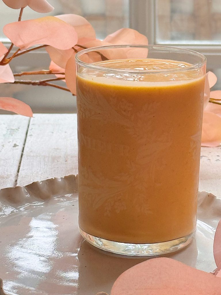 A glass filled with orange smoothie sits on a pale ceramic plate, with pink decorative leaves in the background.