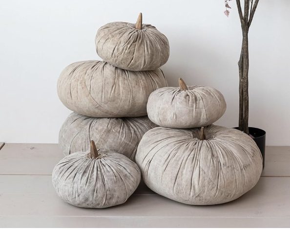 Five fabric pumpkins in neutral tones are stacked and arranged beside a potted plant on a light-colored floor against a white wall.