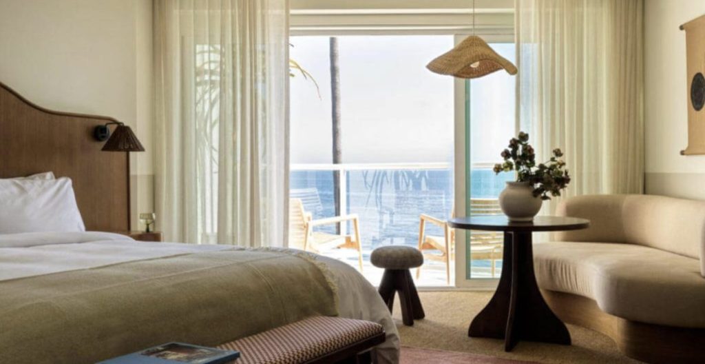 A hotel room with a bed, round table, sofa, and balcony doors open to a view of the ocean; two chairs and a small table are on the balcony.