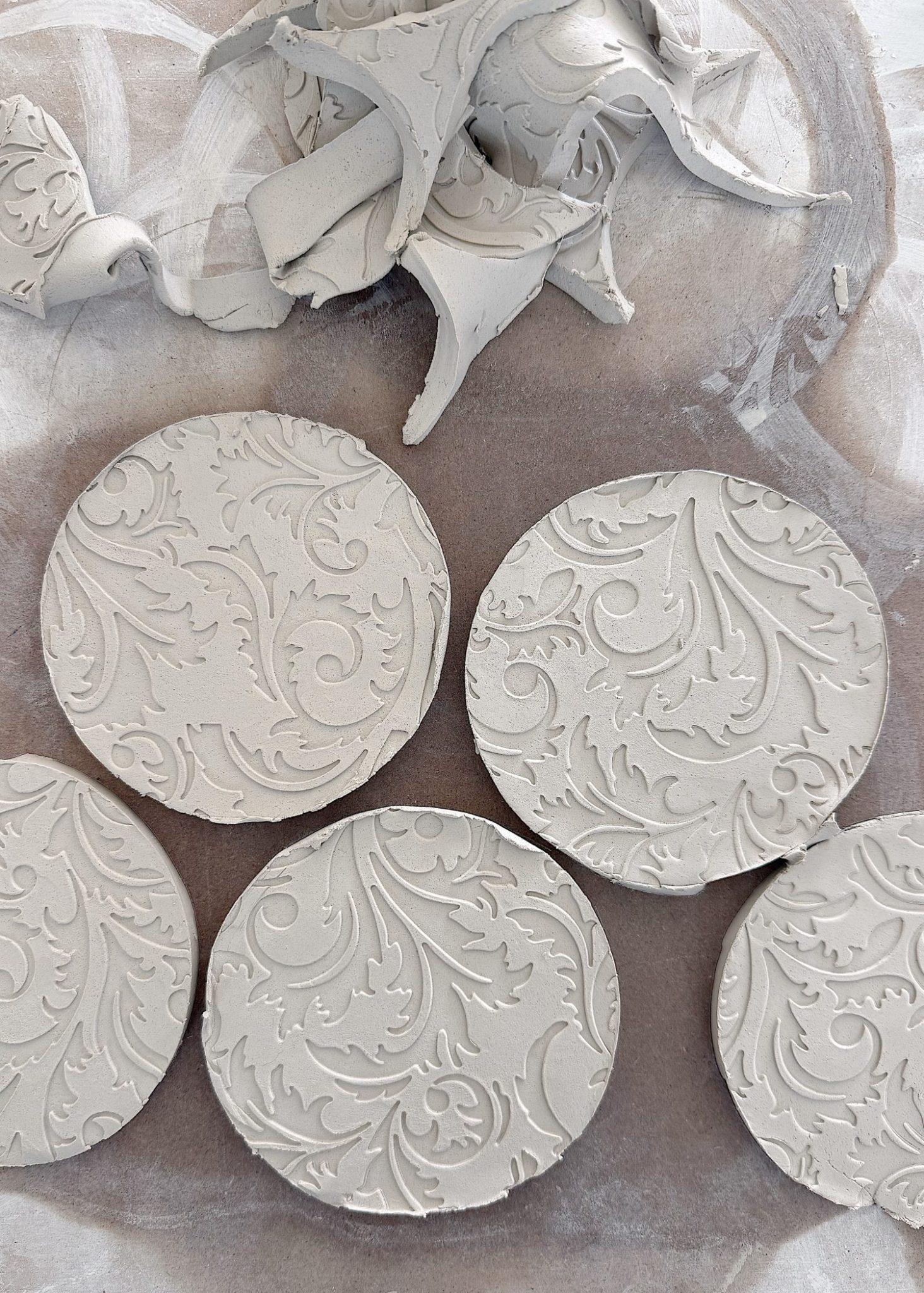 Four round pieces of clay with intricate floral patterns are laid on a surface, with scraps of clay in the background.