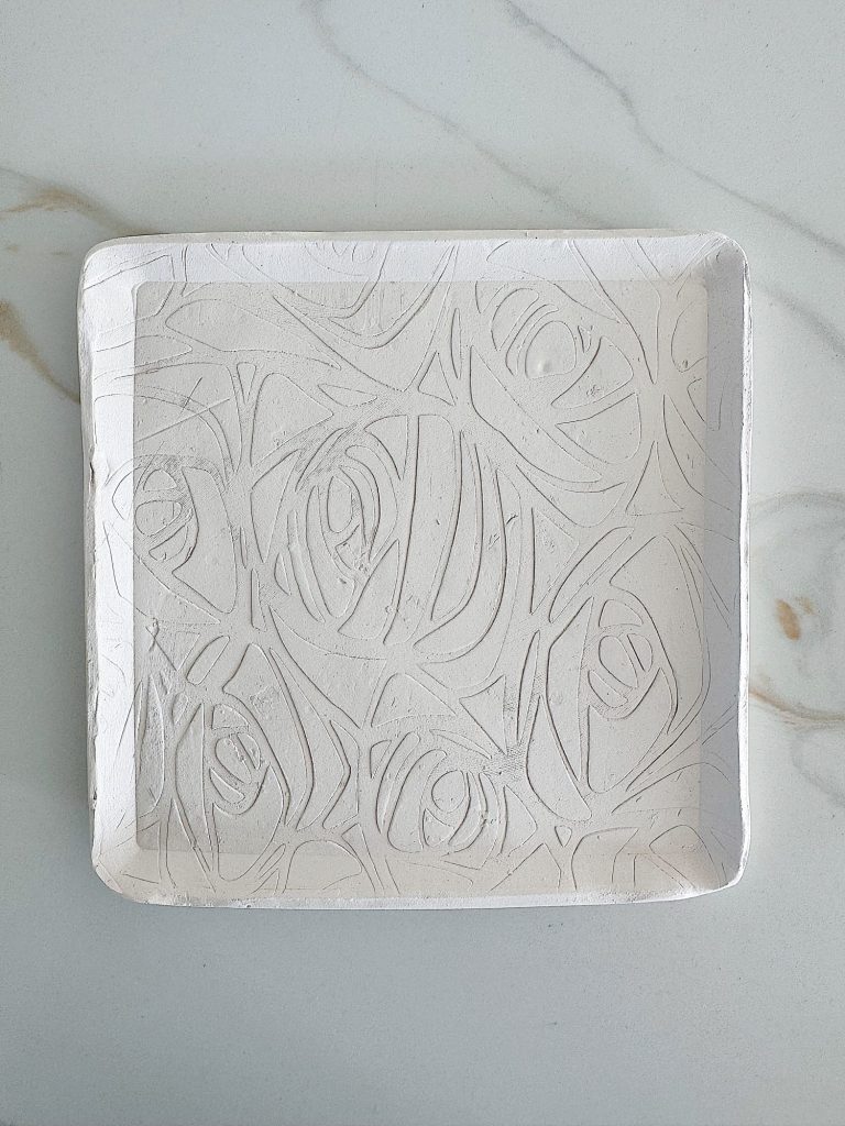 A square white tray with a raised abstract swirl pattern, placed on a light gray marble surface.