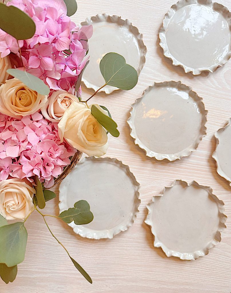 A bouquet of pink hydrangeas and cream roses with green leaves next to seven round, white ceramic plates with wavy edges on a light wooden surface.