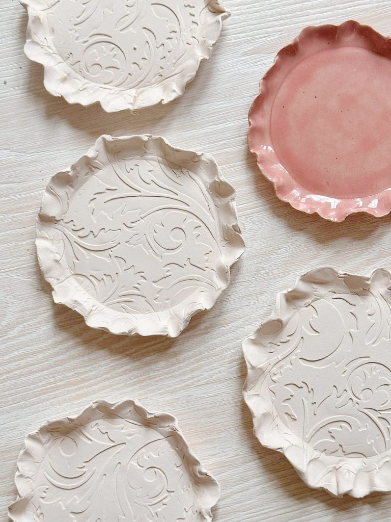 Five decorative ceramic plates with wavy edges are arranged on a light wooden surface; one plate is pink, and the others are white with embossed patterns.