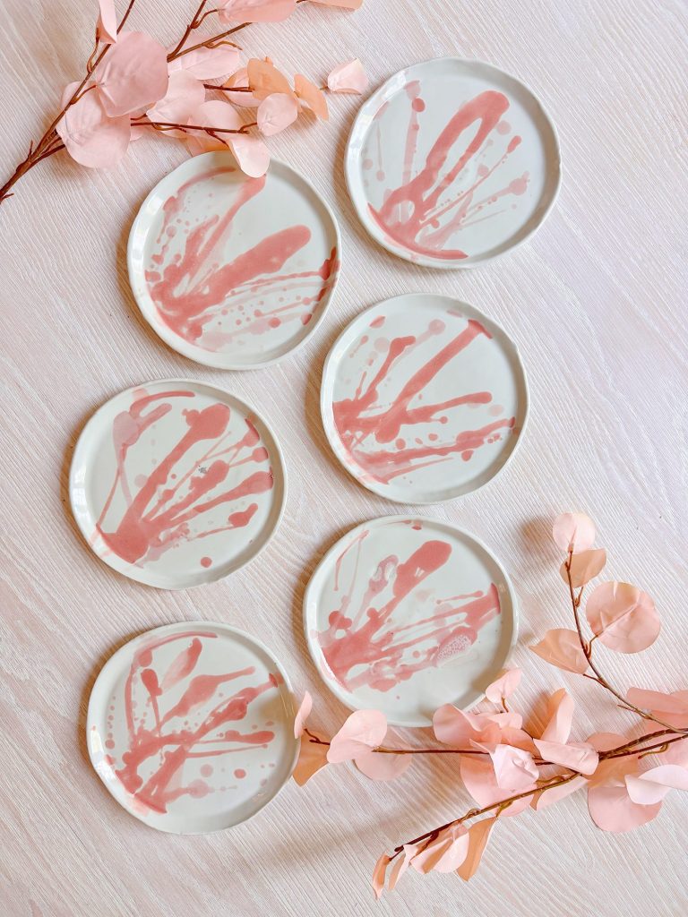 Six round white plates with abstract pink splatter designs are arranged on a light wooden surface, accompanied by pink artificial branches.