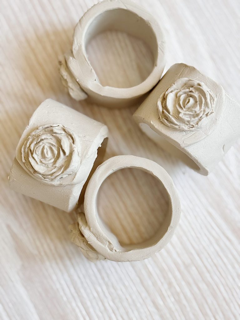 Three beige ceramic napkin rings with floral rose designs are arranged on a light wooden surface.