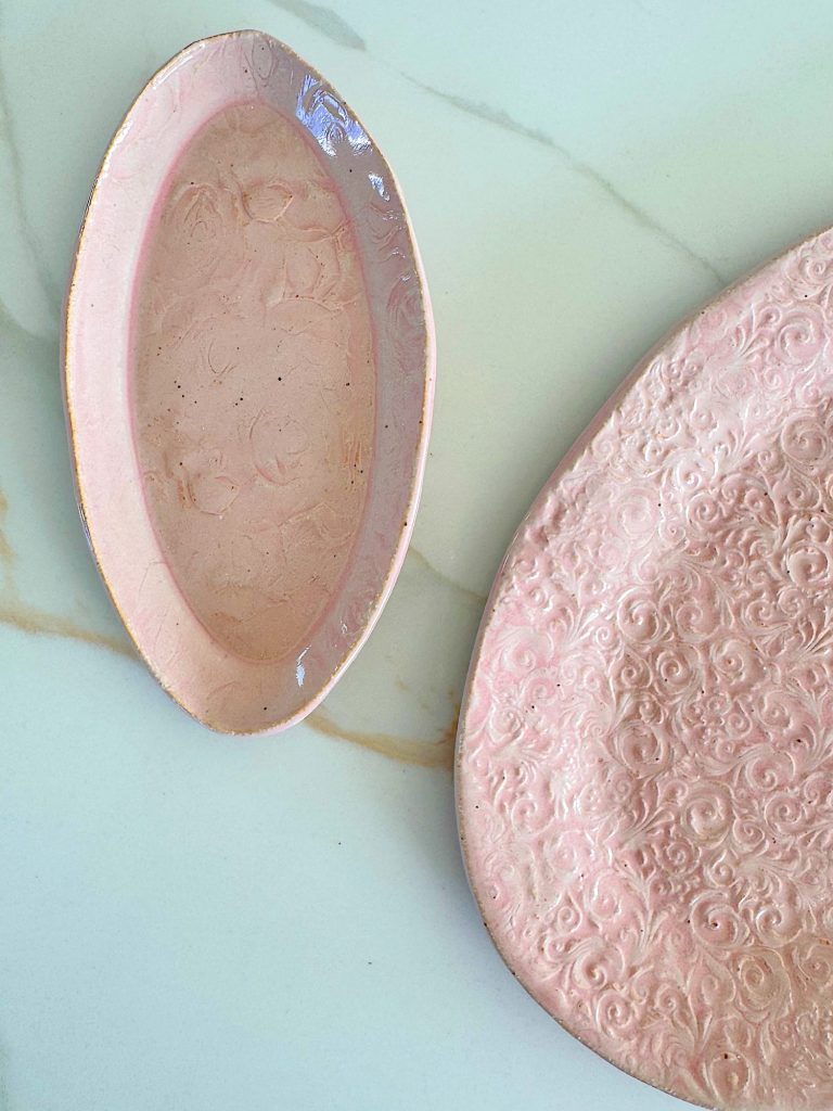 Two light pink ceramic plates with embossed floral patterns on a white marble surface. One plate is oval-shaped, the other is round and partially visible.