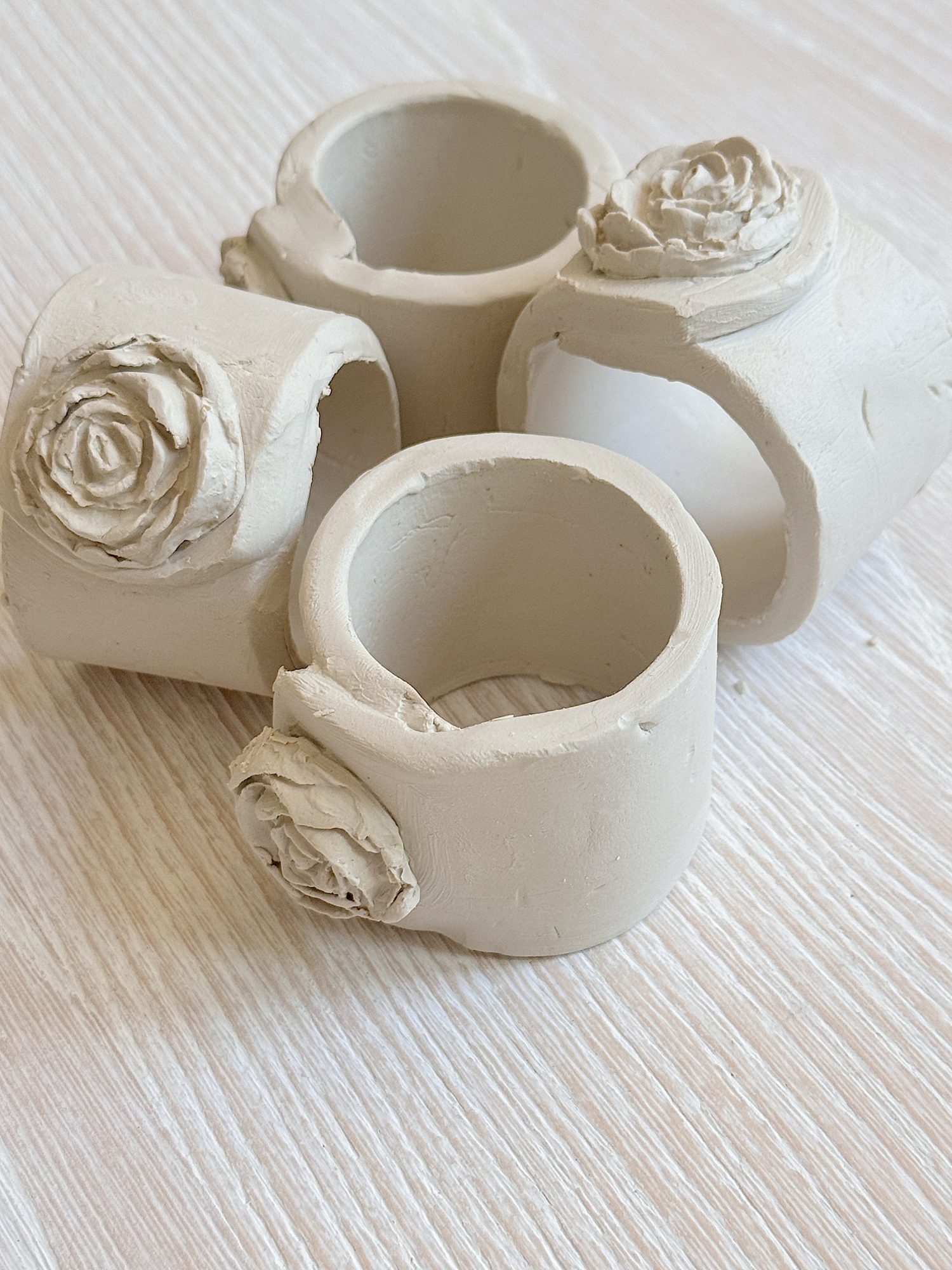 Four unfinished clay napkin rings with rose-shaped decorations are arranged on a light wood surface.