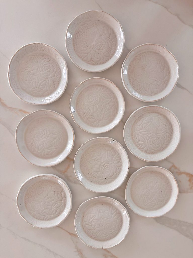 Ten white ceramic plates with embossed floral patterns are arranged in a triangle shape on a light-colored marble surface.