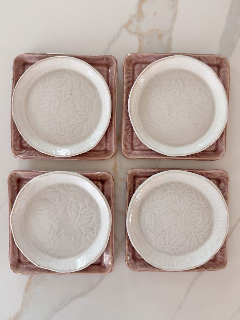 Four white ceramic plates with a floral pattern are placed on four square pink ceramic saucers, arranged in a grid on a light marble surface.