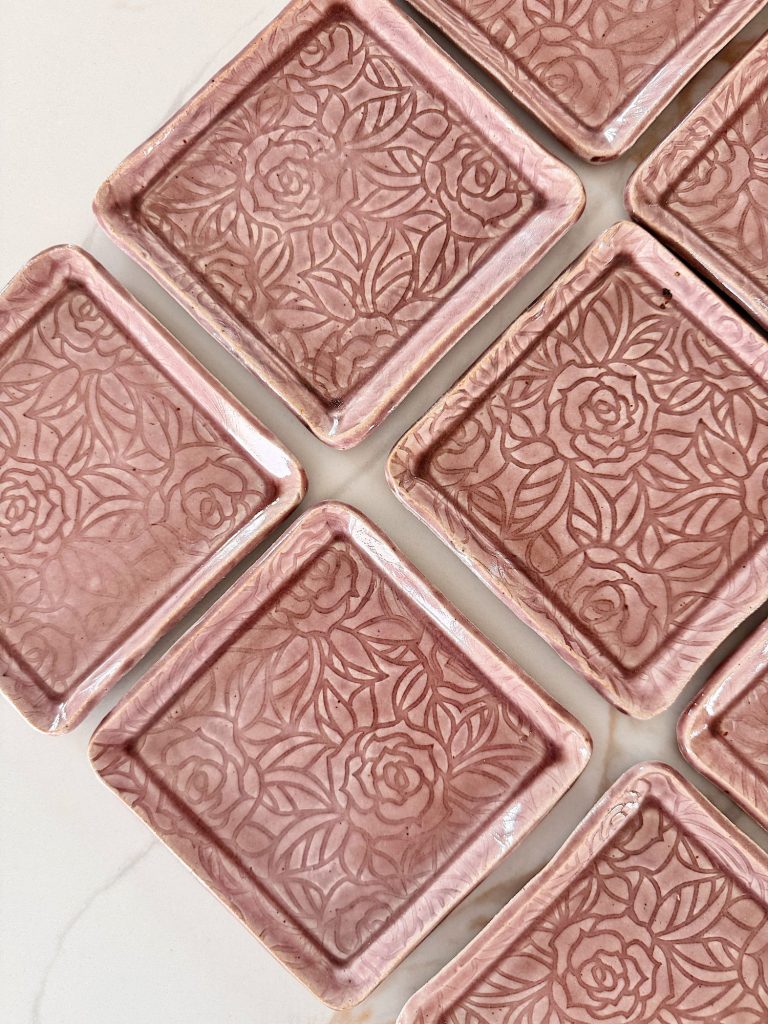 Several pink ceramic square coasters with a raised floral pattern are arranged in a diagonal layout on a light surface.