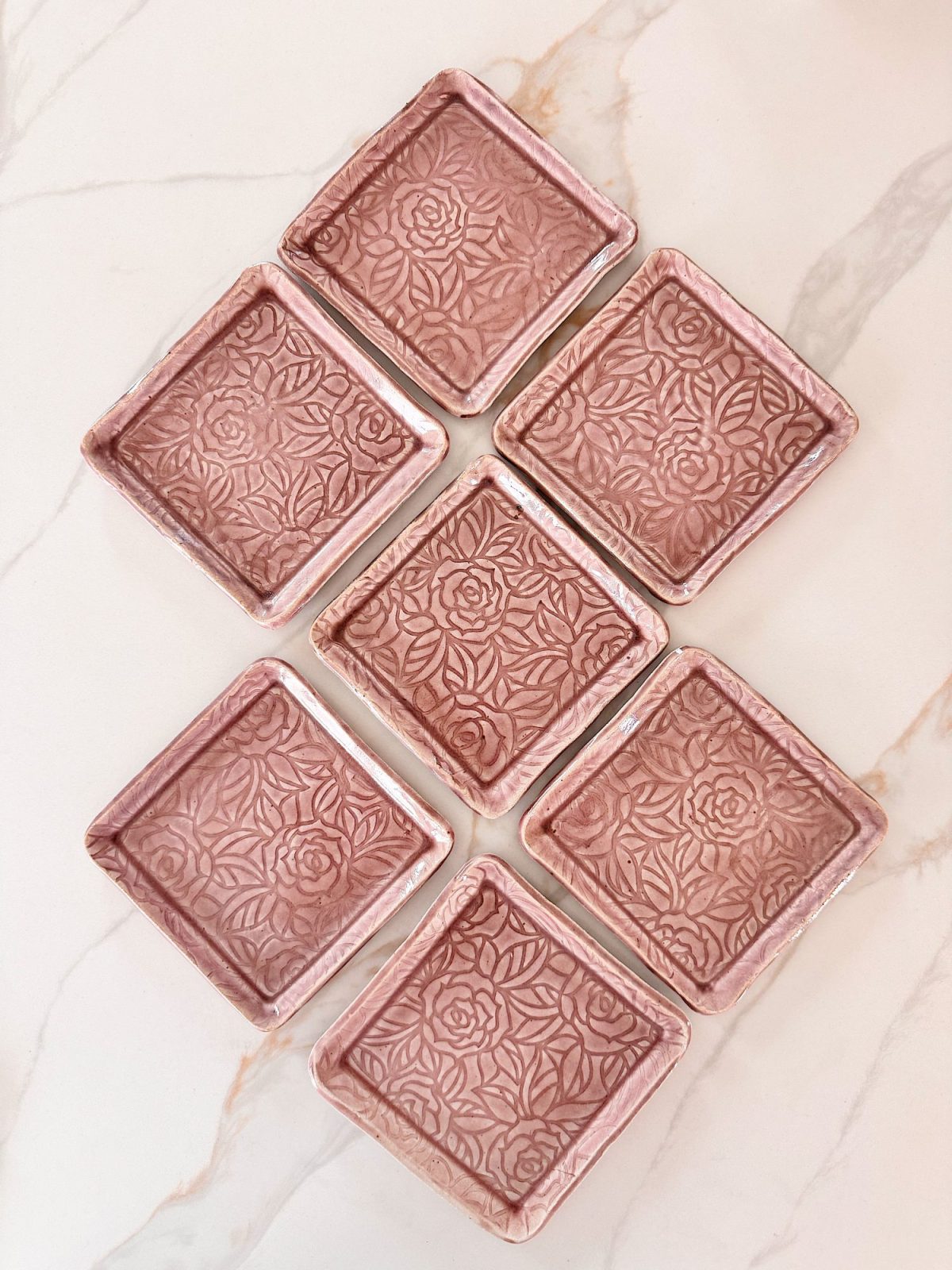 Seven pink square ceramic plates with a floral embossed pattern are arranged in a diamond shape on a light marble surface.