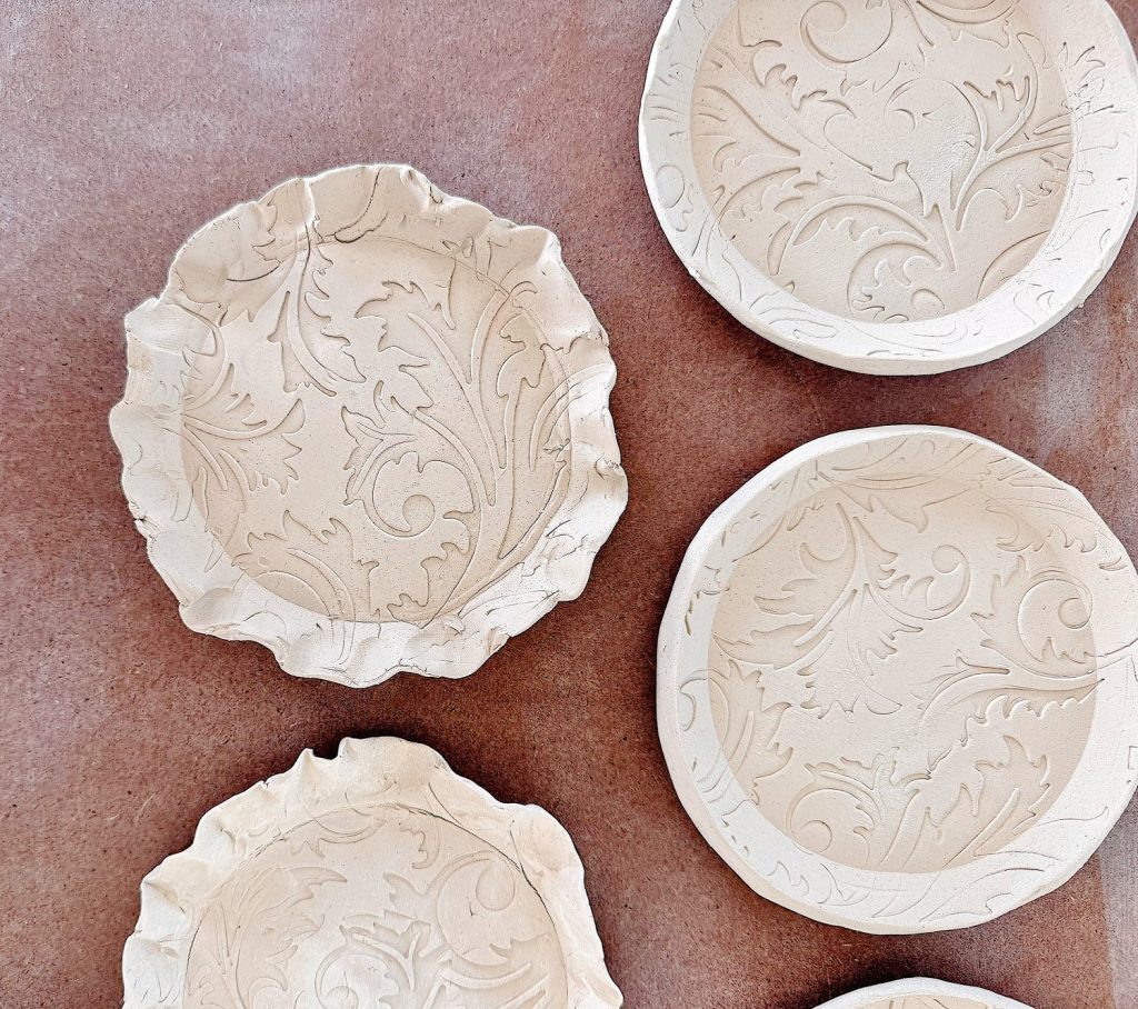 Five unglazed ceramic plates with embossed leaf patterns are arranged on a brown surface. Two plates have ruffled edges, and three plates have smooth, round rims.