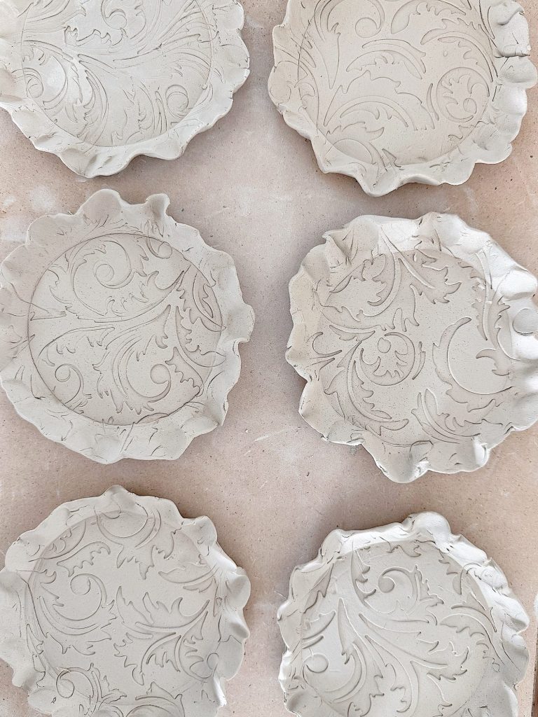 Six unglazed ceramic plates with embossed floral and leaf patterns are arranged on a light surface.