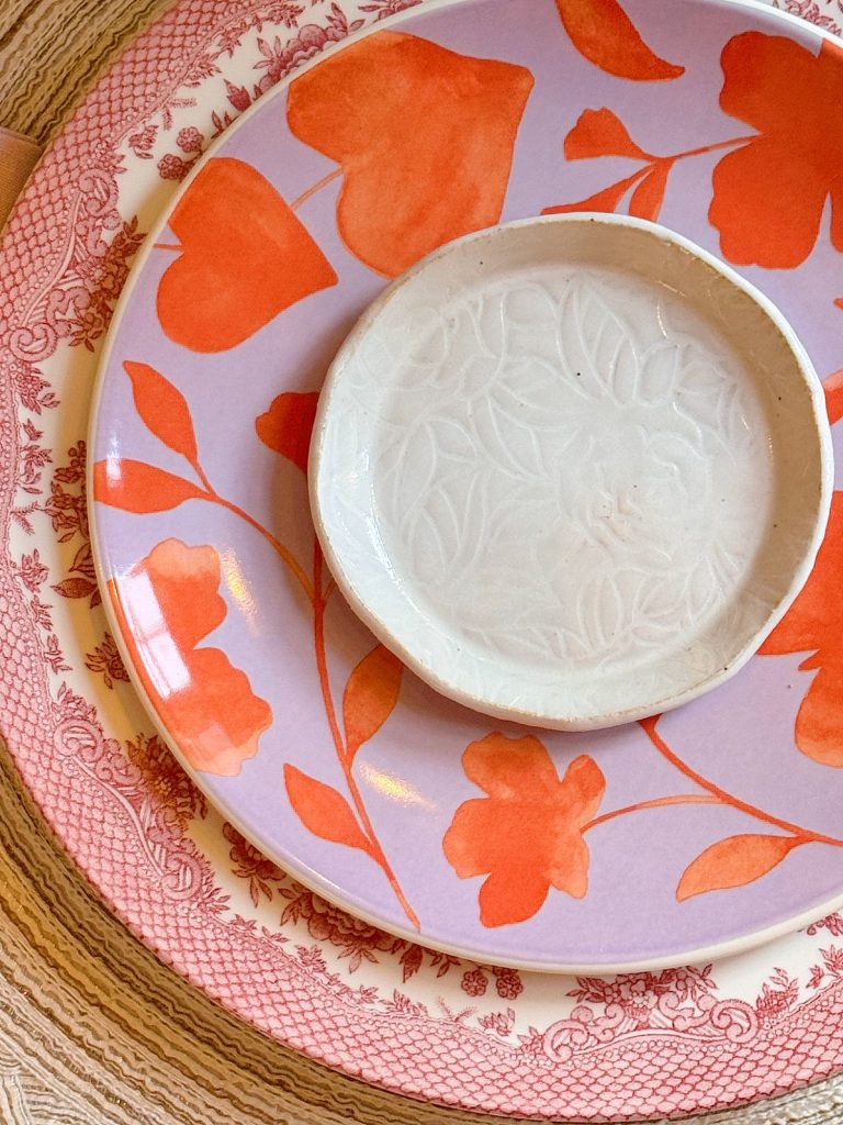 A stack of three plates with varied floral patterns and colors, including a small white dish on top of pink and orange decorative plates set on a woven placemat.