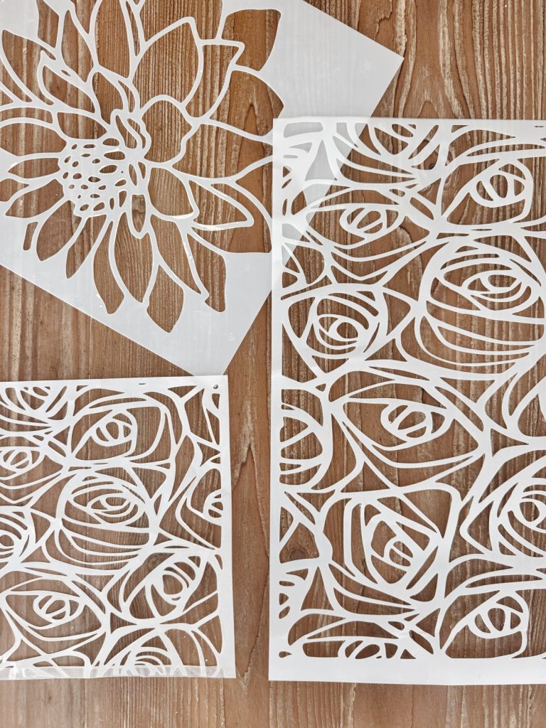 Three white floral-patterned stencils are laid out on a wooden surface; two feature abstract rose designs, and one depicts a single large flower.