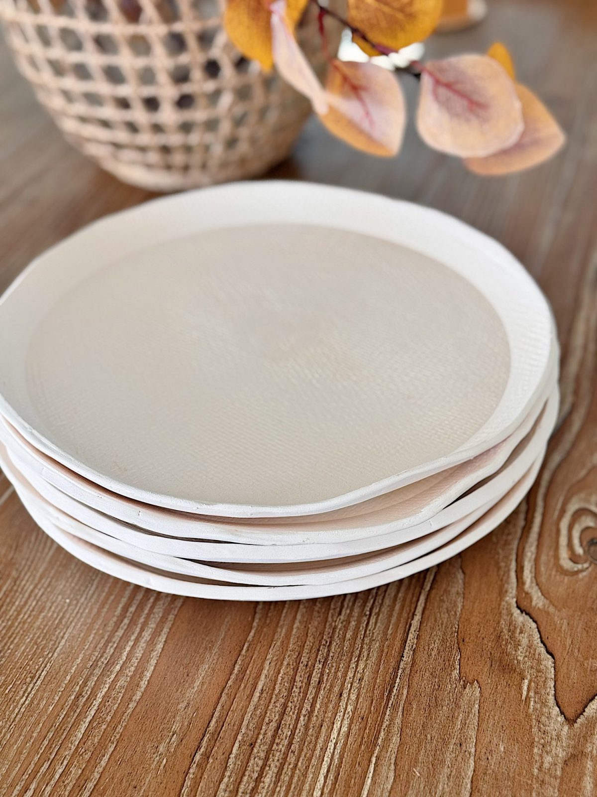 A stack of six white ceramic plates sits on a wooden table, with a woven basket and autumn-colored leaves in the background.