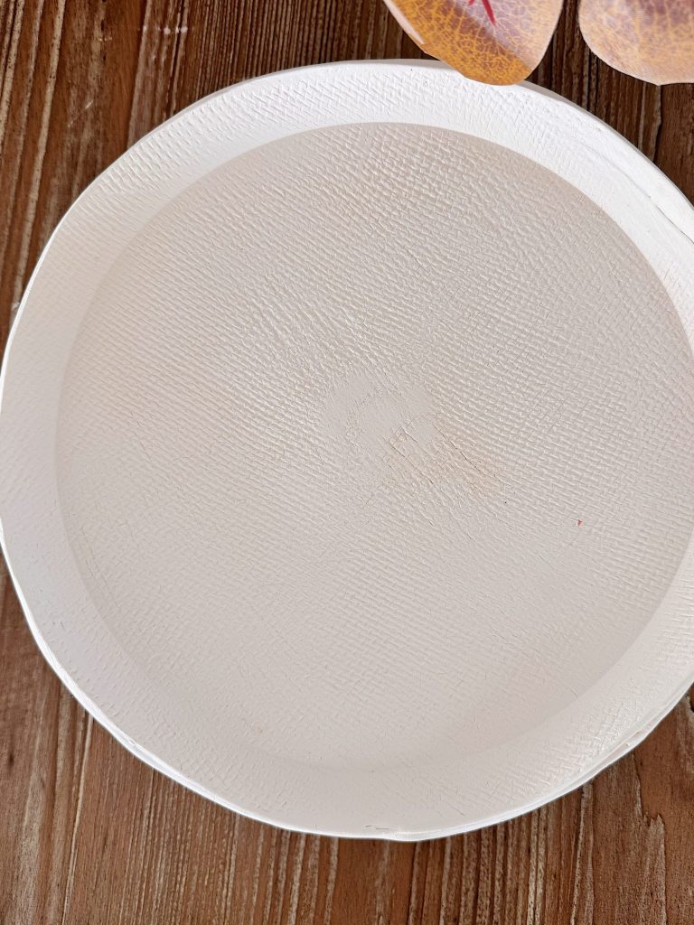A plain white textured paper plate on a wooden surface, with part of a patterned object visible in the top right corner.