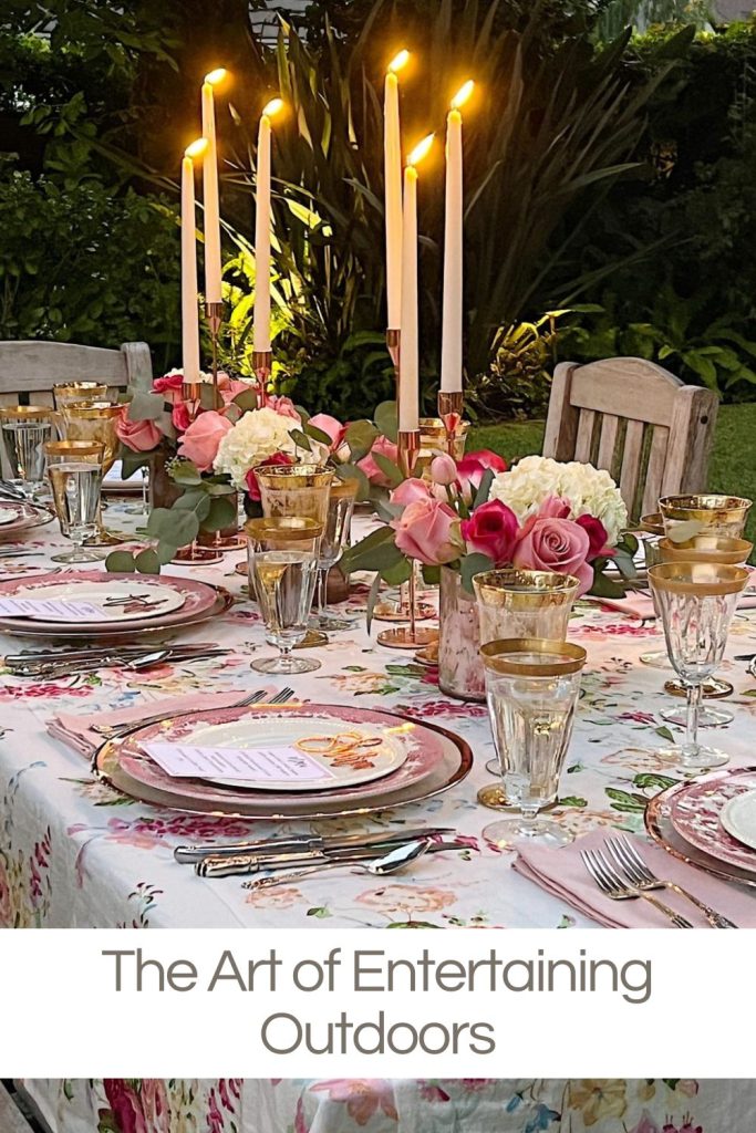 An outdoor dining table is set with floral tablecloth, gold-rimmed glassware, pink flowers, and lit tall candlesticks. Text at the bottom reads, "The Art of Entertaining Outdoors.