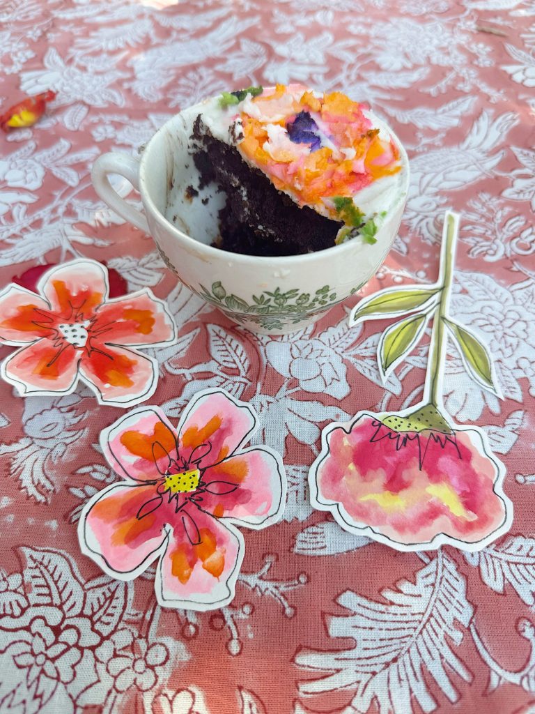 A teacup holding a slice of chocolate cake with colorful frosting sits on a floral tablecloth beside three watercolor-style flower cutouts.