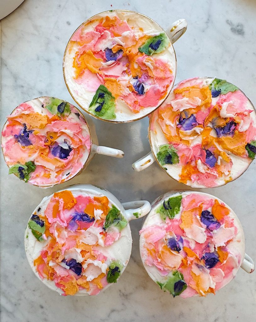 Five cups of latte with colorful, floral-inspired latte art on a marble surface, featuring pink, orange, green, and purple decorative designs on the foam.