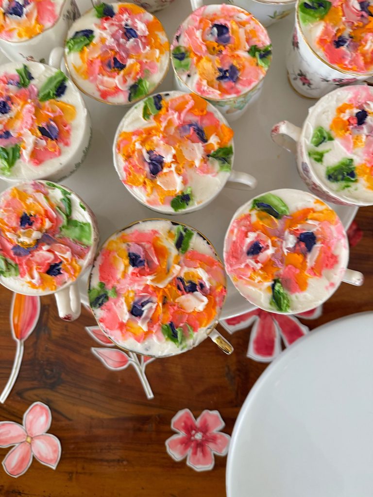 Several teacups filled with a creamy dessert topped with colorful, floral-inspired decorations are arranged on a wooden table next to pink flower-shaped coasters and a white plate.