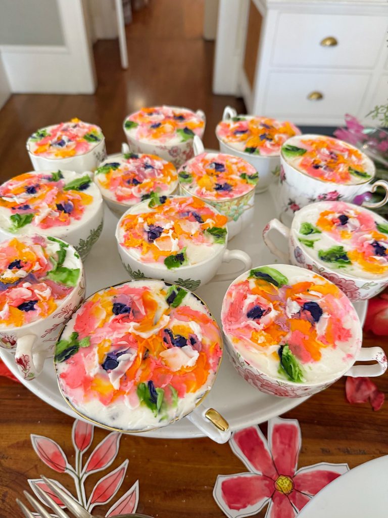 Teacups filled with a dessert topped with colorful frosting flowers, arranged on a white platter on a wooden table.