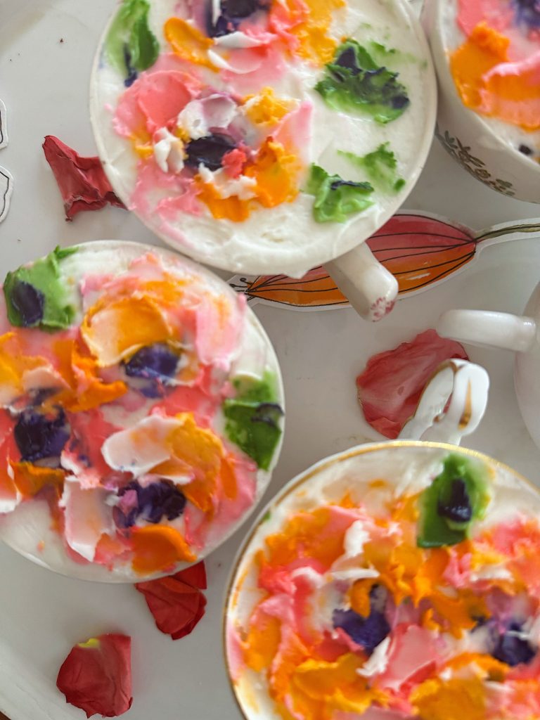 Three cups filled with a creamy white substance topped with colorful flower petal-like decorations, placed on a white surface with scattered red and pink petals.