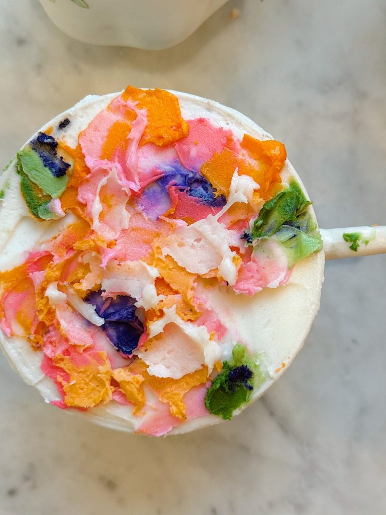 A cup of coffee topped with a layer of colorful, textured foam or cream in shades of pink, orange, green, purple, and white on a marble surface.