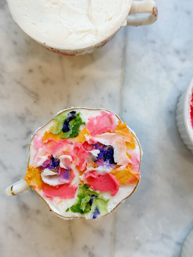 A teacup filled with a drink topped with colorful, flower-like icing or cream sits on a marble surface next to another cup and a white bowl.